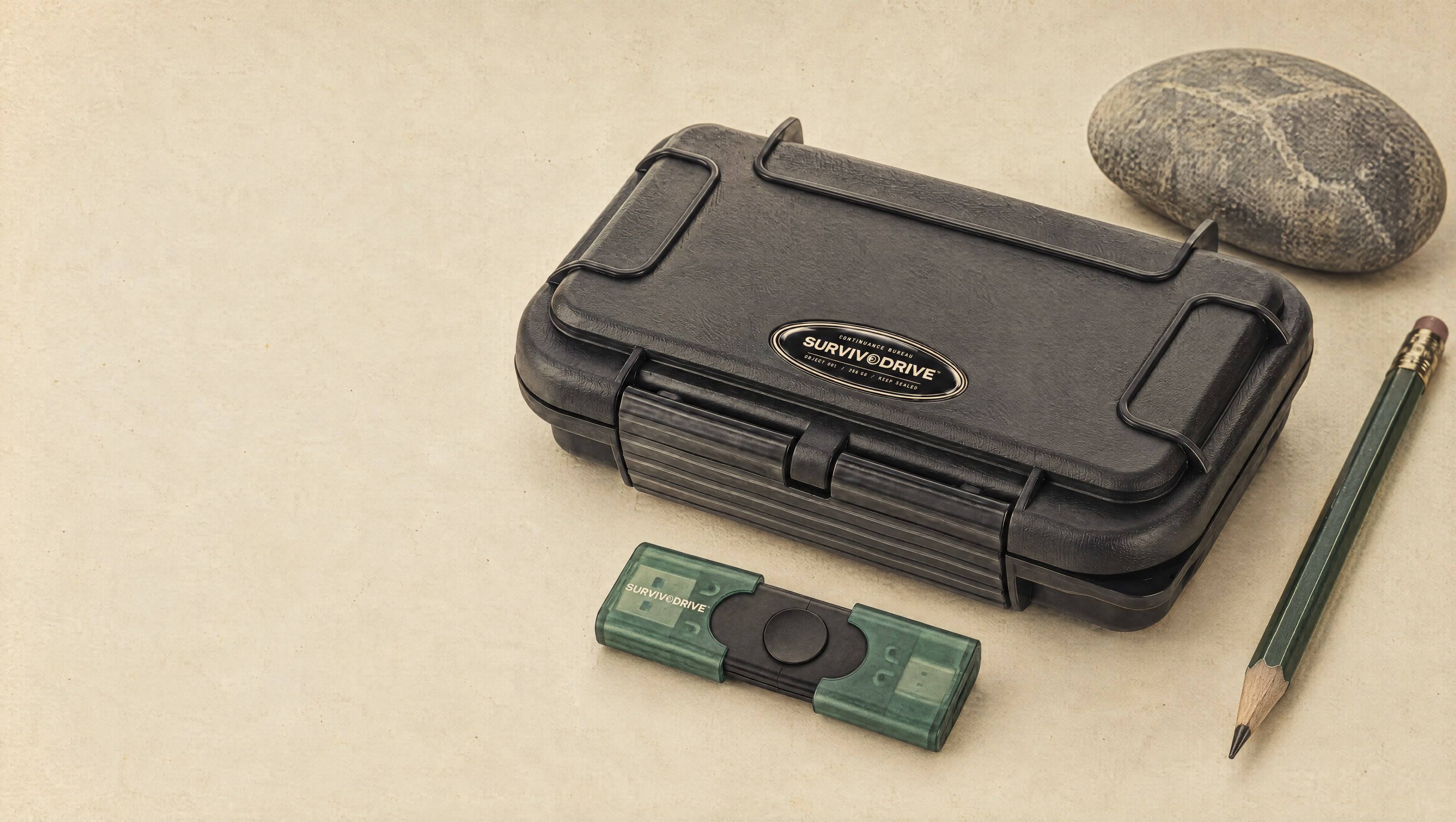 SURVIVODRIVE: the black waterproof travel case with its domed oval badge, the dual USB drive with translucent caps, a river stone and a pencil on bone-coloured paper